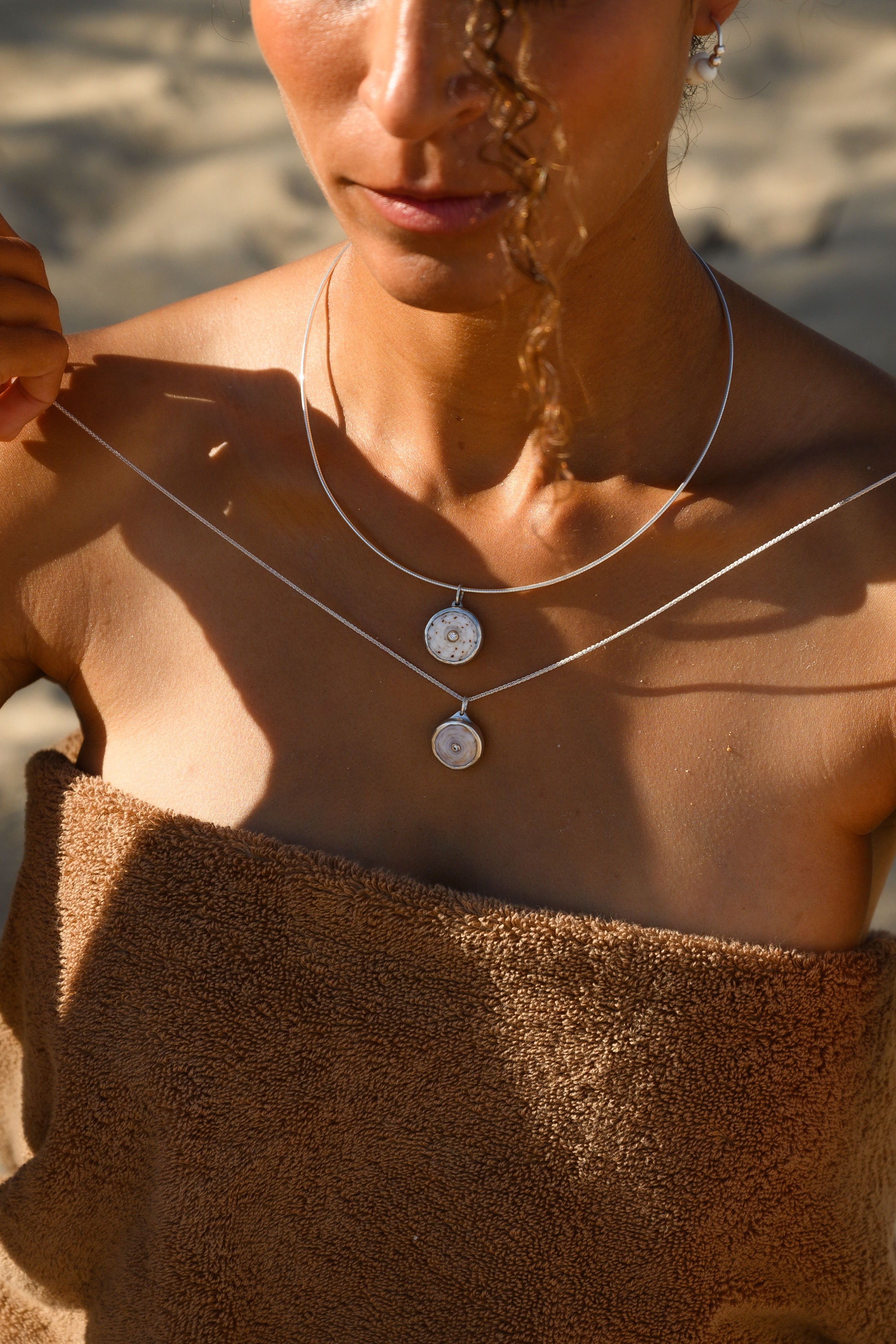 Person wearing a silver necklace with two circular pendants on a beach.